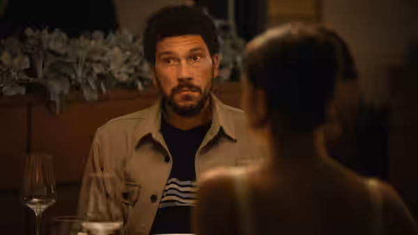 A bearded man in a jacket looks across a table at a woman in a restaurant