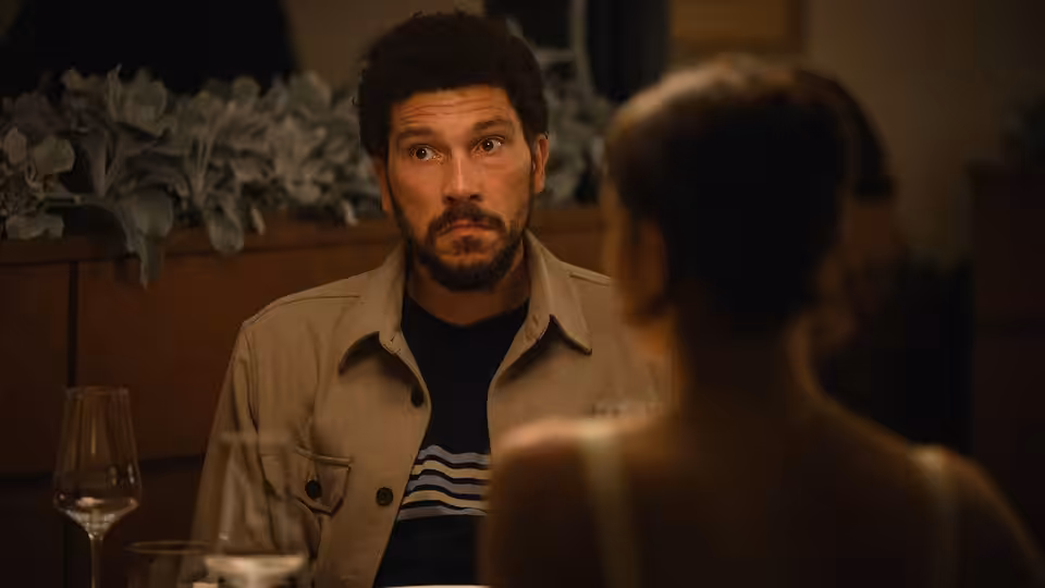 A bearded man in a jacket looks across a table at a woman in a restaurant