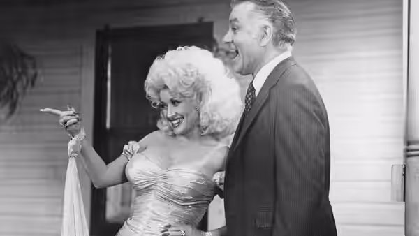 Mona Stangley (Dolly Parton) pointing as a man in a suit stands beside her on a porch