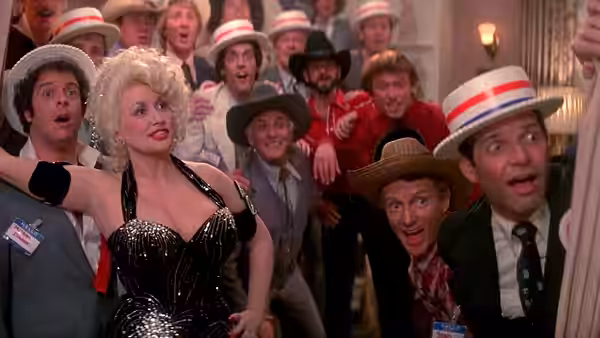 Mona Stangley (Dolly Parton) among a group of surprised men in a crowded room