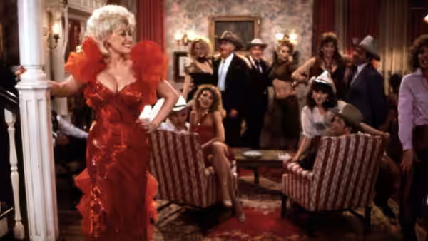 Mona Stangley (Dolly Parton) standing in a red dress inside a busy parlor