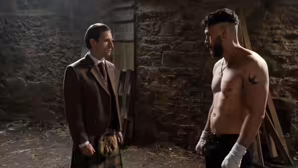 Niall in a kilt faces shirtless Ruben in a stone cellar