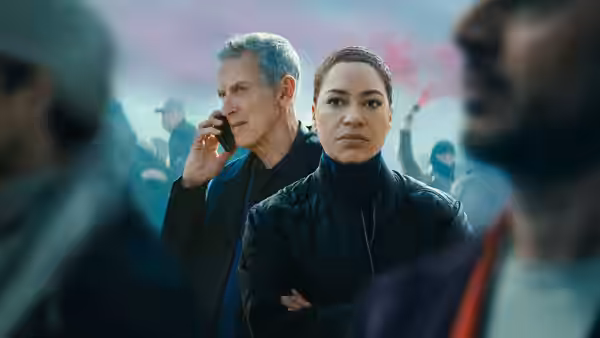 DCI Daniel Hegarty (Peter Capaldi) on the phone next to DS June Lenker (Cush Jumbo) in a crowd