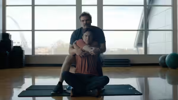 David Harbour hugging a man seated on a yoga mat in a gym
