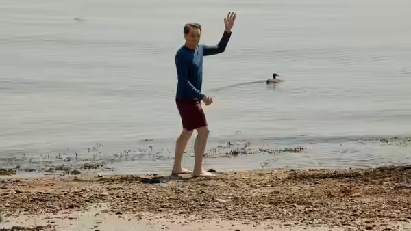 a man waves while standing on the shore of a calm bay