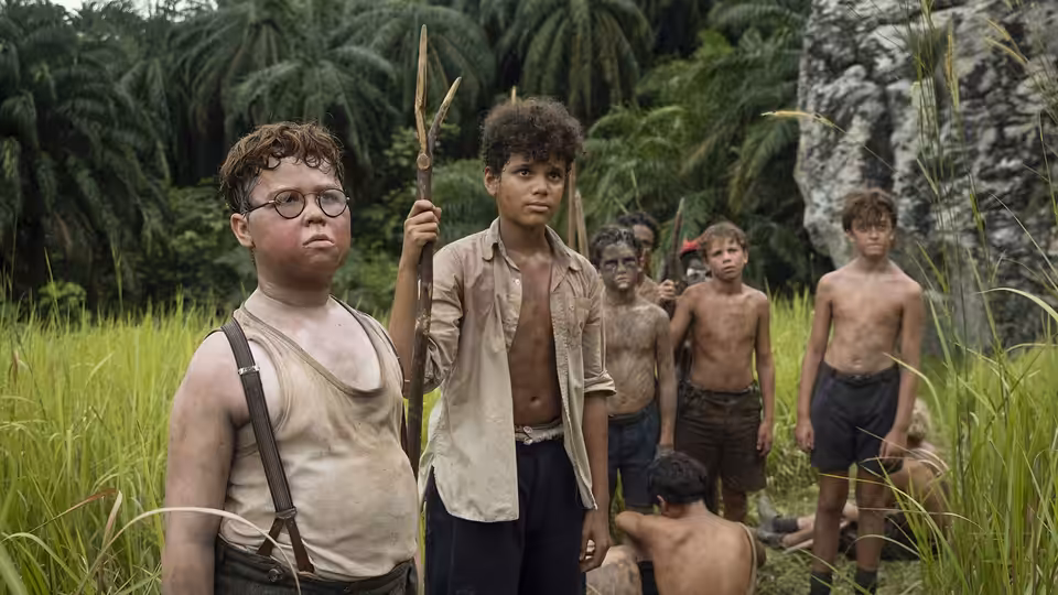 A boy with glasses stands next to one holding a spear among others in grass