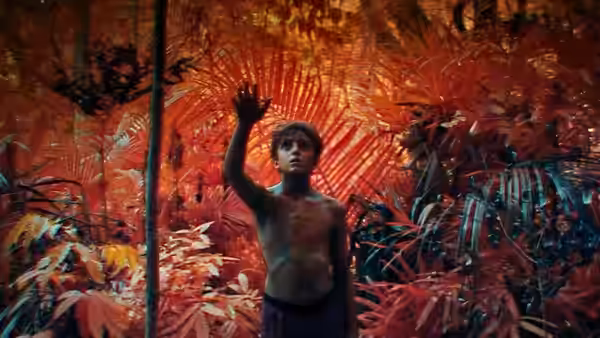 A shirtless boy stands in dense red jungle foliage with one arm raised