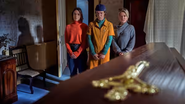 Three people standing in a room with a coffin in the foreground