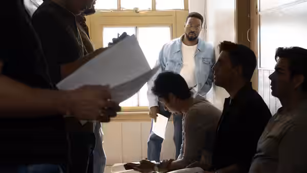Simon Williams (Yahya Abdul-Mateen II) standing behind men reviewing papers indoors
