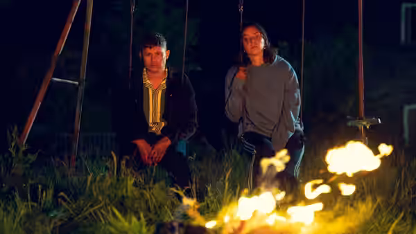 Young man and woman sit on swings by a campfire at night