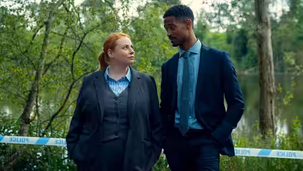 Red haired woman and suited man stand near police tape by a lake