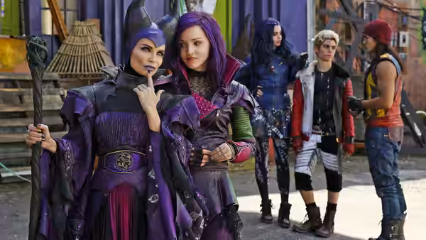 Mal (Dove Cameron) and Evie (Sofia Carson) stand in front of purple and blue buildings