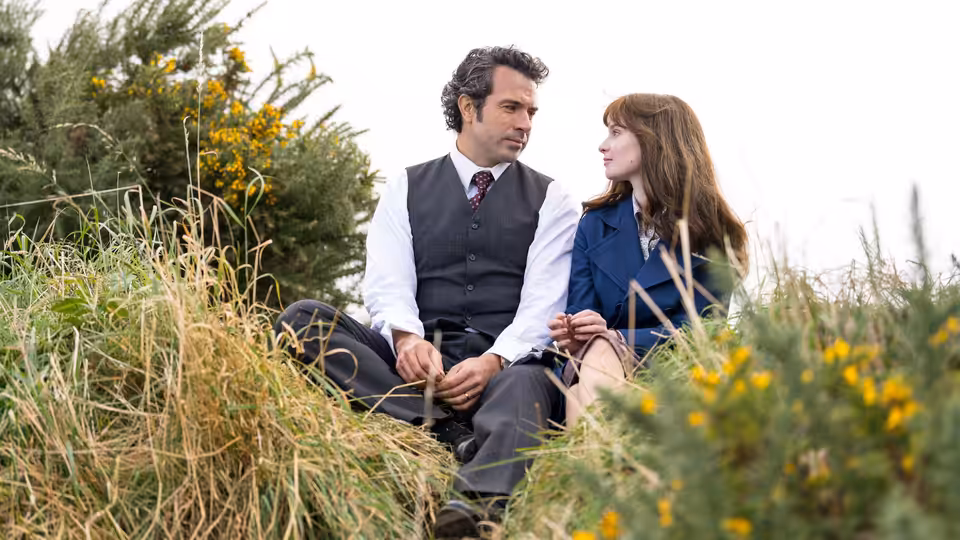 A man and a woman sit closely together on a grassy hillside
