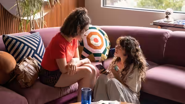 Two women sit close on a purple couch while one holds a phone