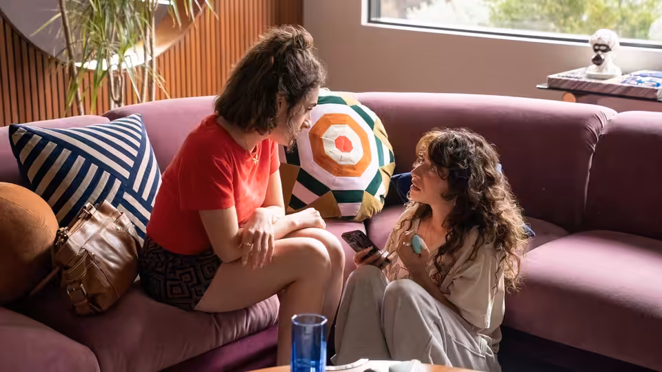 Two women sit close on a purple couch while one holds a phone