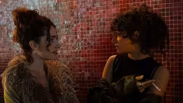 Two women face each other closely before a red tiled wall