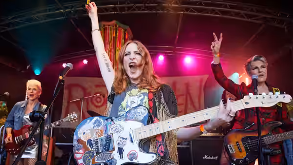 Red-haired guitarist raises arm while singing on stage with two bandmates