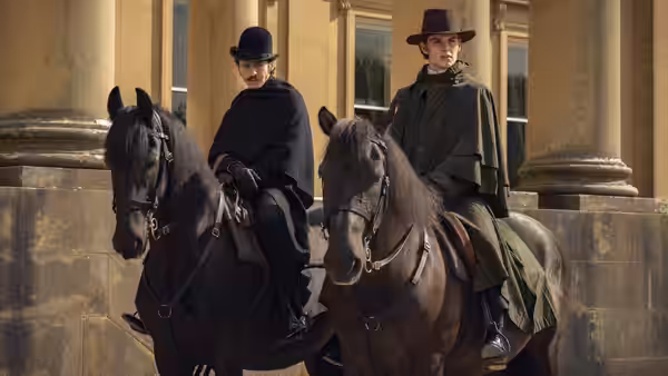 Two men in period clothing riding horses in front of a large building