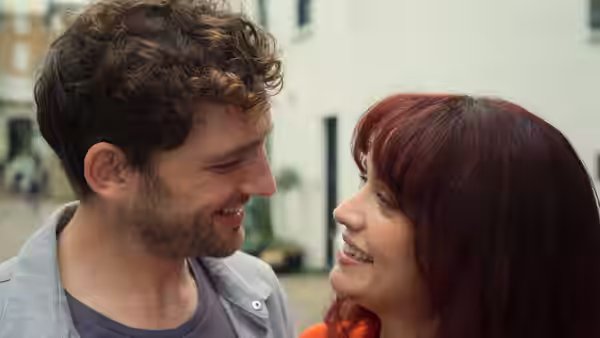 Curly-haired man and red-haired woman smiling at each other outdoors