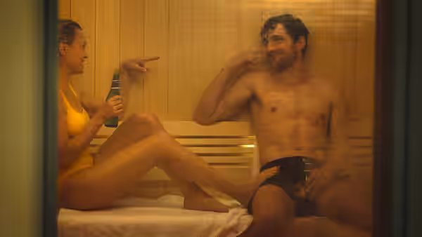 Woman in yellow swimsuit pointing at shirtless man inside wooden sauna
