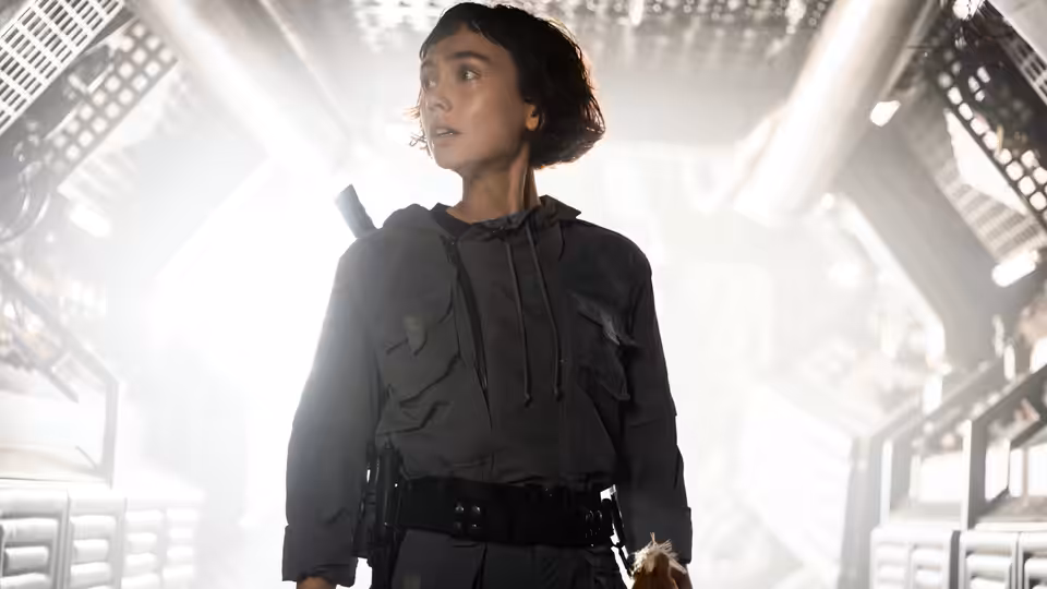 Wendy (Sydney Chandler) standing alert in a brightly lit industrial corridor