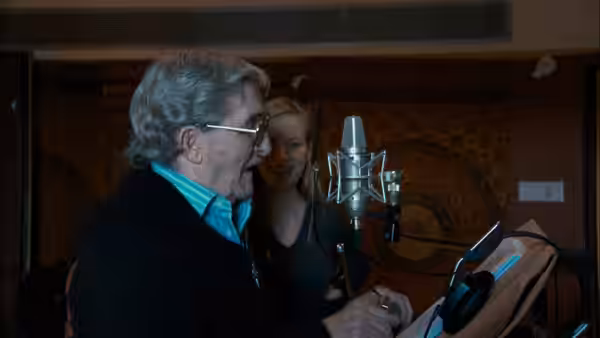 an older man speaking into a studio microphone with a woman nearby
