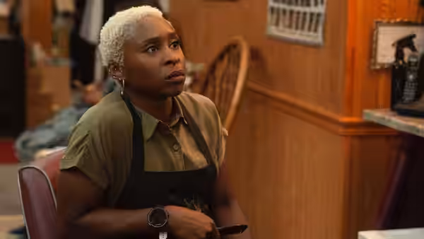 Cynthia Erivo as Belle seated in a shop wearing an apron