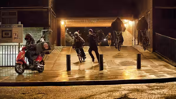 A group of youths on bicycles and a scooter ride through a housing estate at night