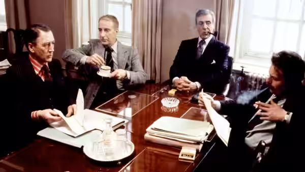 Four men in suits seated around a conference table with papers and ashtray