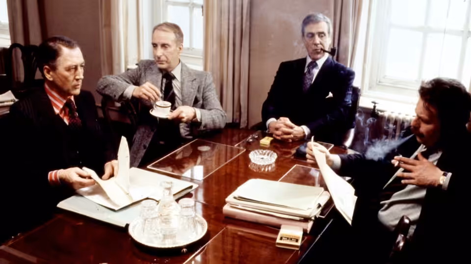Four men in suits seated around a conference table with papers and ashtray