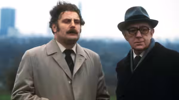 Two men in overcoats stand outdoors with a blurred cityscape behind them