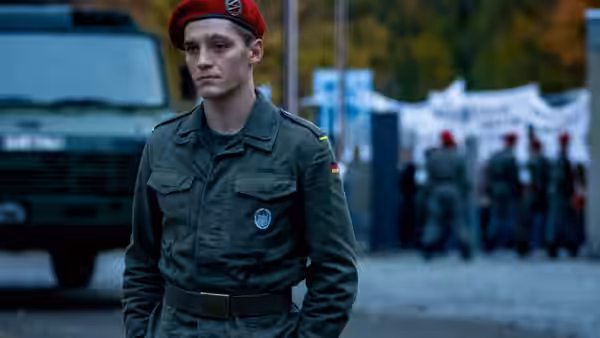 Soldier in red beret stands before a military vehicle with troops behind