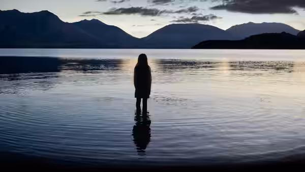 woman standing in calm lake at dusk with mountains behind