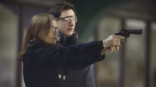 Michèle (Isabelle Huppert) aiming a revolver with a man behind her