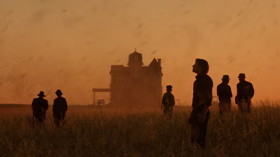 Men in hats stand in tall grass before a large building at dusk
