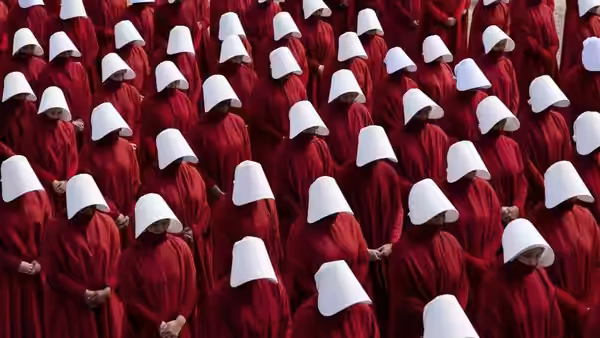 Rows of handmaids in red cloaks and white bonnets standing with heads bowed
