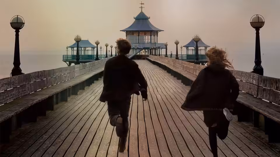A man and a woman running along a wooden pier towards a pavilion