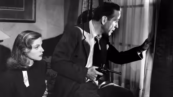 Vivian (Lauren Bacall) sits beside Philip Marlowe (Humphrey Bogart) who holds a gun and peers out curtains