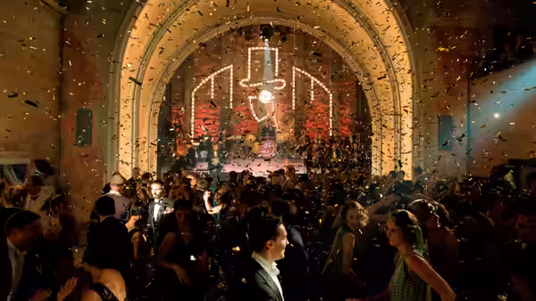 Crowd dances under falling confetti in a grand hall with glowing neon sign