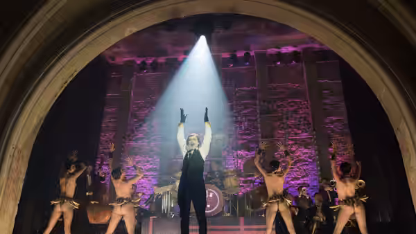 Man in tuxedo raises arms center stage amid shirtless dancers and a band