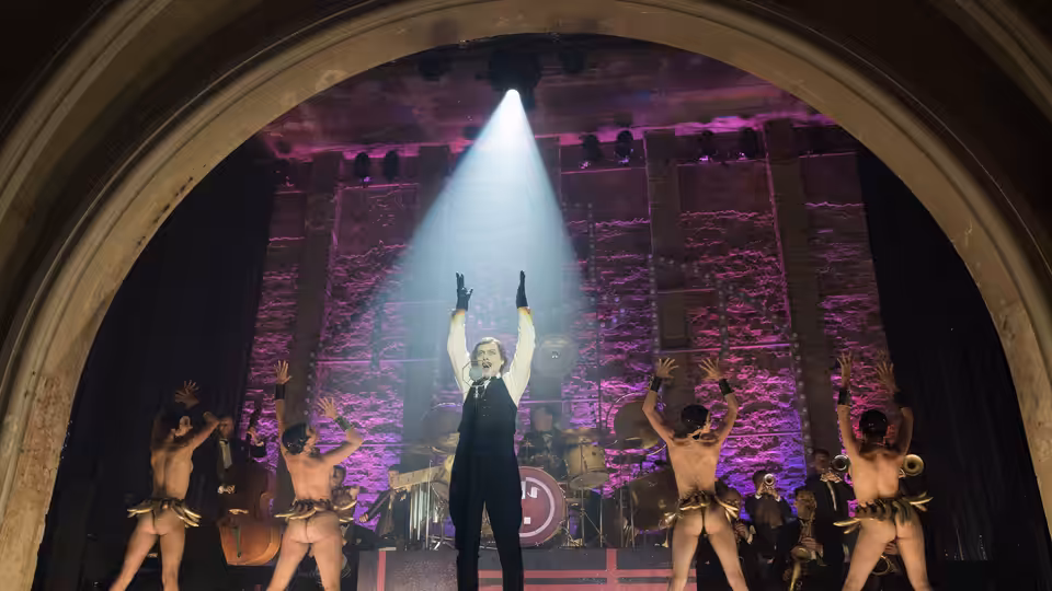 Man in tuxedo raises arms center stage amid shirtless dancers and a band