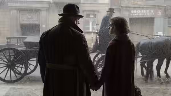 Man and woman in coats hold hands in the rain beside horse carriages