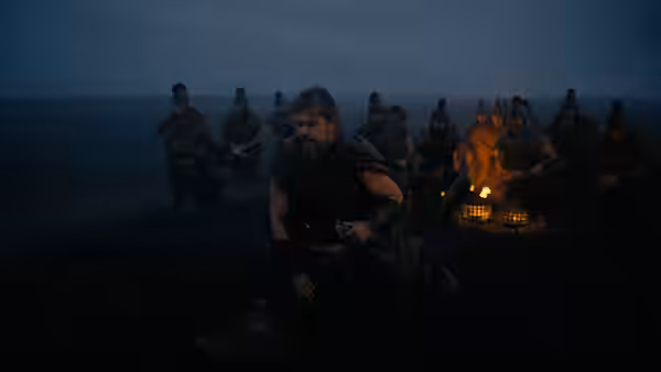 A bearded warrior crouches by a campfire surrounded by seated men at dusk