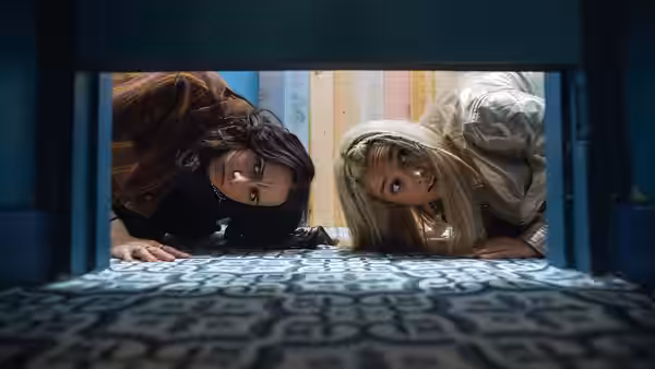Josie (Kat Sadler) and Billie (Lizzie Davidson) lie on patterned carpet peering through an open doorway