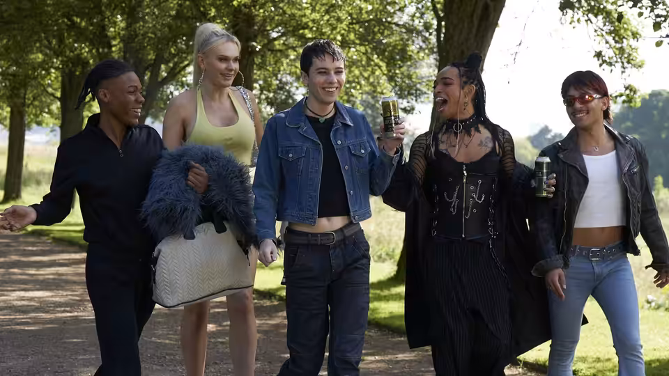 Five friends in bold outfits walk a wooded path holding drinks and bags