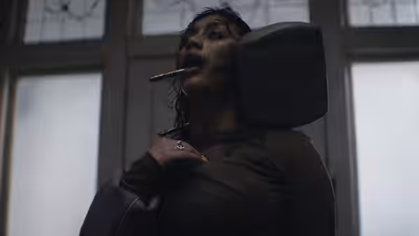 A bloodied woman with a pencil in her mouth clutches her chest indoors