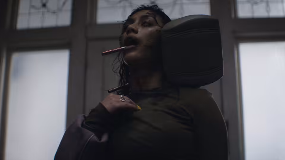 A bloodied woman with a pencil in her mouth clutches her chest indoors