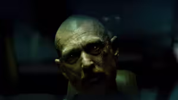 A dirty-faced bald man with a beard stares ahead in dim light