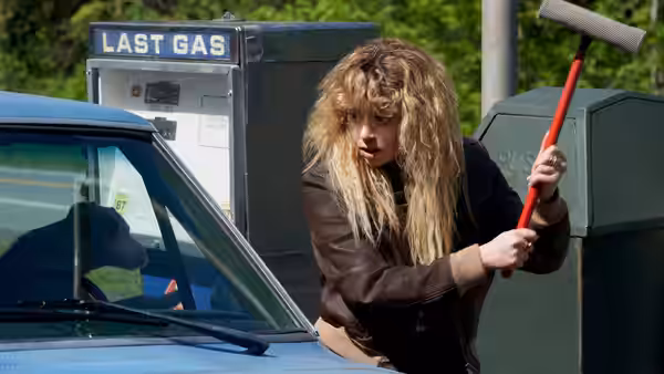 a woman with blonde hair holding a sledgehammer beside a blue car at a gas station