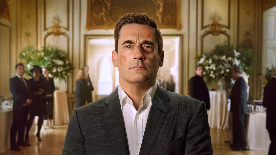 Coop (Jon Hamm) standing in an ornate hall with guests at tables